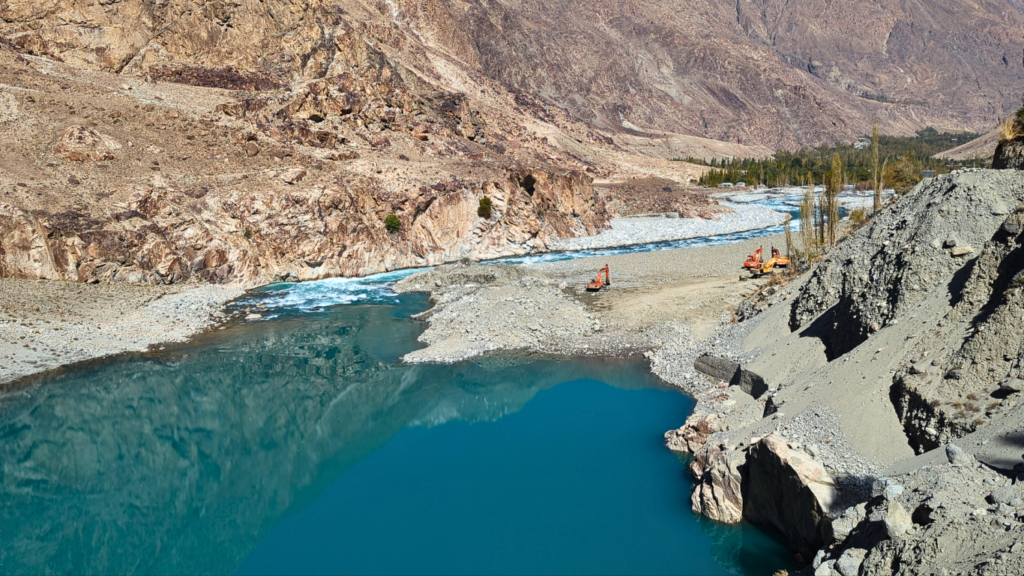 Blocked Ghizer River after the August 2025 GLOF in Talidass, showing a debris dam, turquoise floodwater, and excavators clearing sediment in a mountain valley.